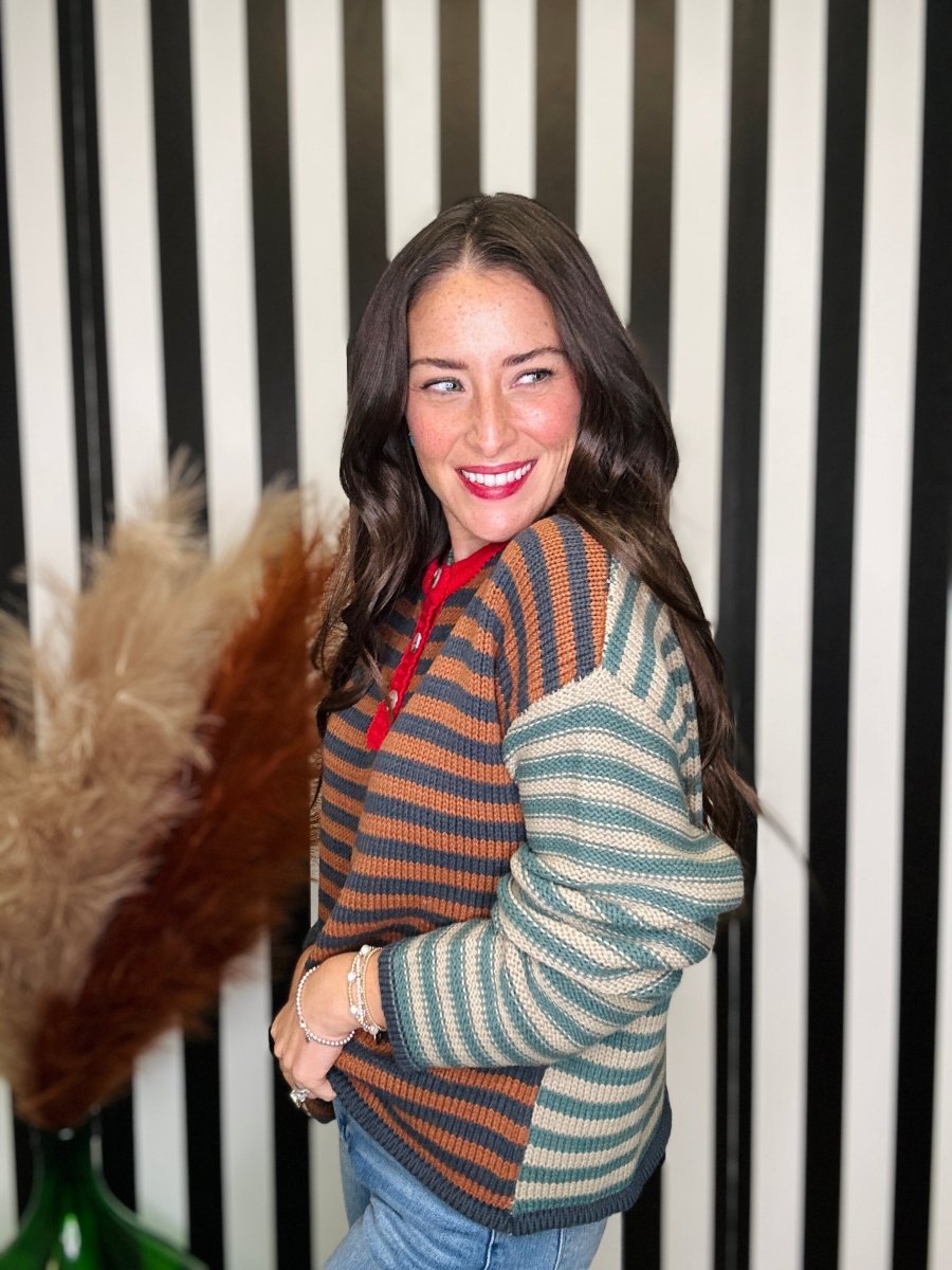 A woman with long dark hair smiling and looking over her shoulder, wearing a color-block knit sweater with horizontal stripes in rust orange, teal green, and cream, paired with blue jeans. She is standing in front of a black and white vertically striped wall.