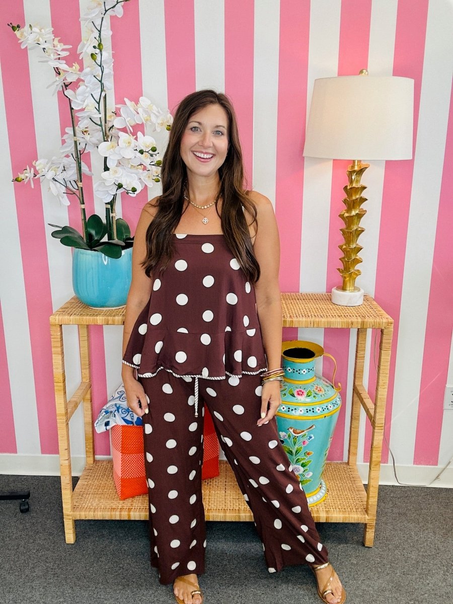 A woman modeling a brown two-piece pajama set with white polka dots, consisting of a sleeveless top and matching pants, standing in front of a pink and white striped wall.