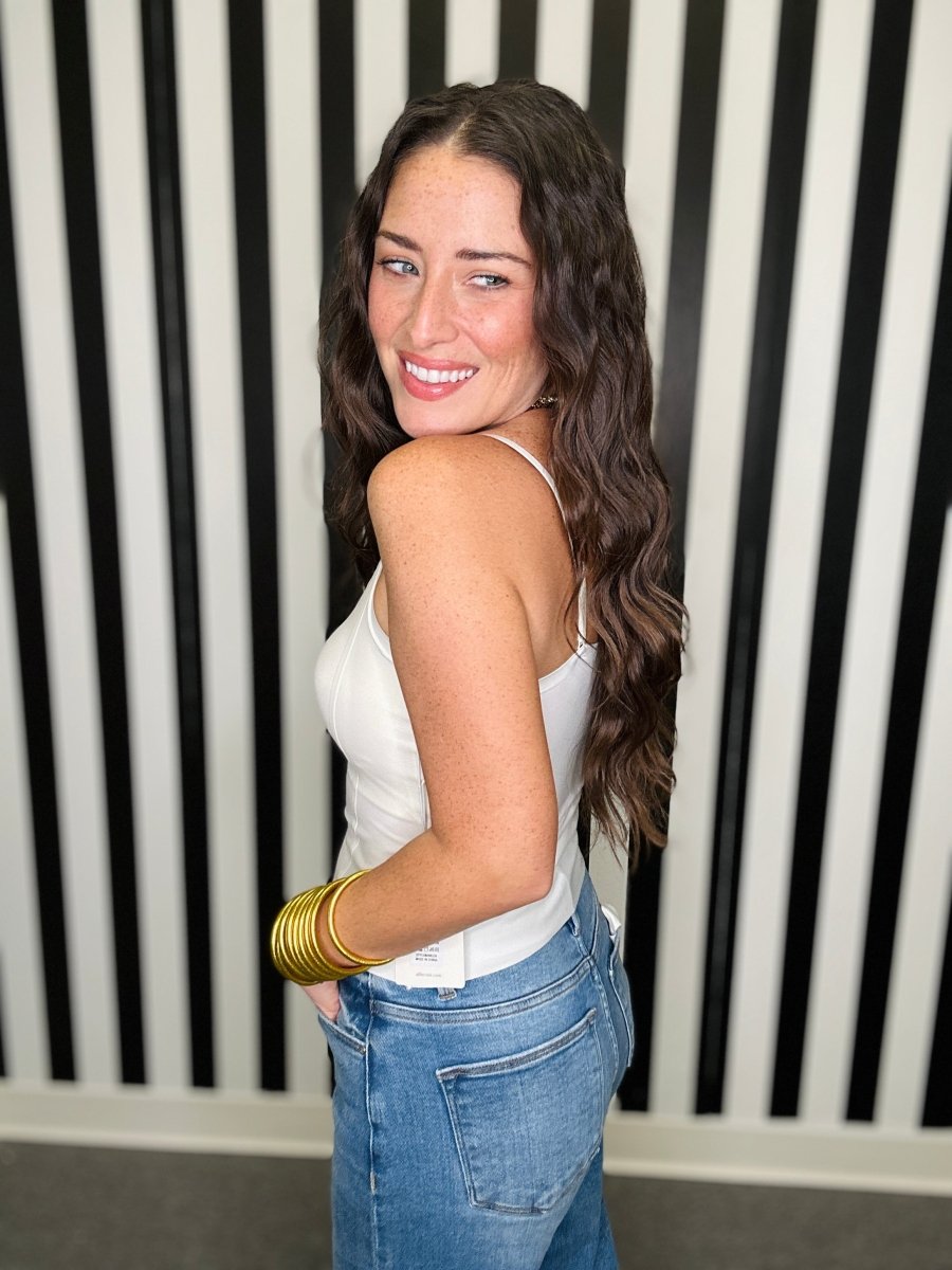 A woman with long wavy brown hair is shown from the back-three-quarter view, wearing a white tank top and blue jeans, accessorized with gold bangles, standing in front of a black and white vertically striped wall.