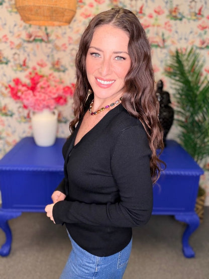 A woman with long wavy brown hair smiling, wearing a black ribbed long-sleeve V-neck top and blue jeans, standing in front of a floral wallpaper background with a blue side table.