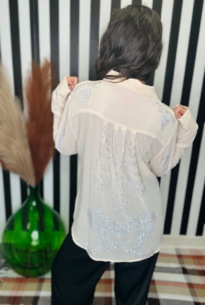A person viewed from behind wearing a sheer cream long-sleeve blouse with silver sequined butterfly patterns on the back, paired with black pants, standing in front of a black and white striped wall.