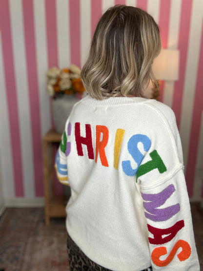 A woman viewed from behind wearing a white knit sweater with the word CHRISTMAS spelled out in large multicolored letters across the back and sleeves. The sweater is shown in a pink-and-white striped room setting.