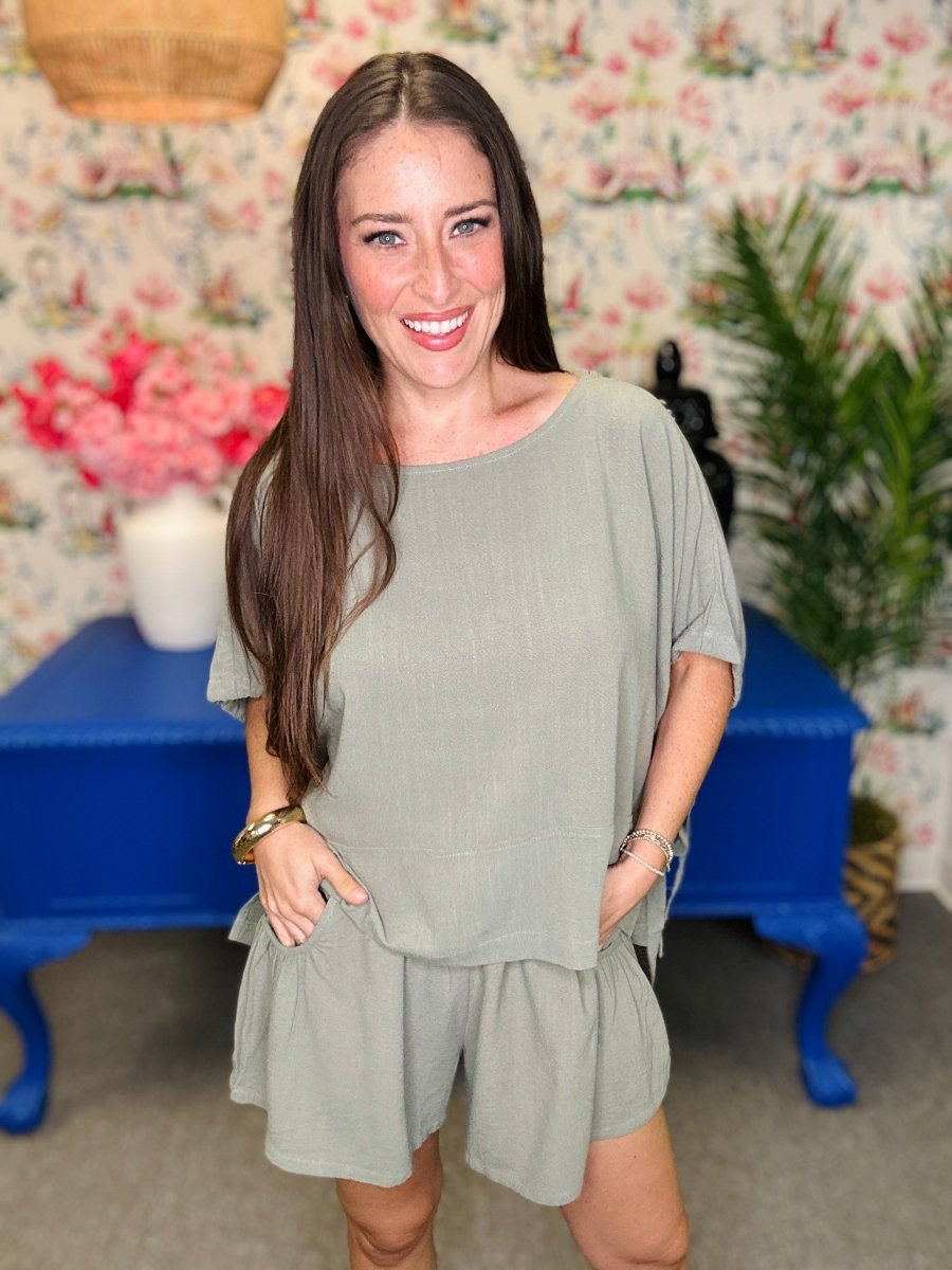 A woman modeling a sage green short-sleeve romper with a relaxed fit, standing in front of a floral wallpaper background with a blue bench.