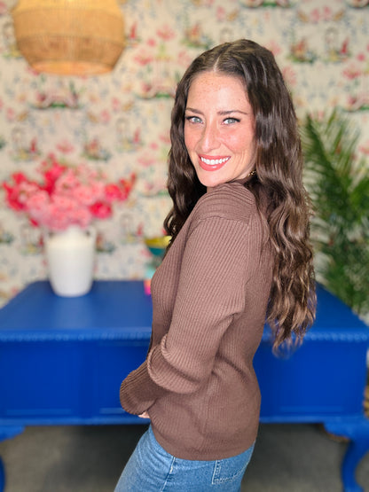 A woman with long wavy brown hair smiles while looking back over her shoulder in a ribbed brown sweater and blue jeans, standing in front of a floral wallpaper background with a blue table.