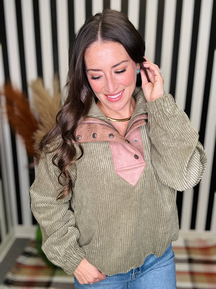 A woman wearing a corduroy pullover sweater with a sage green body and a dusty pink mock neck collar, paired with blue jeans, standing in front of a black and white striped background.