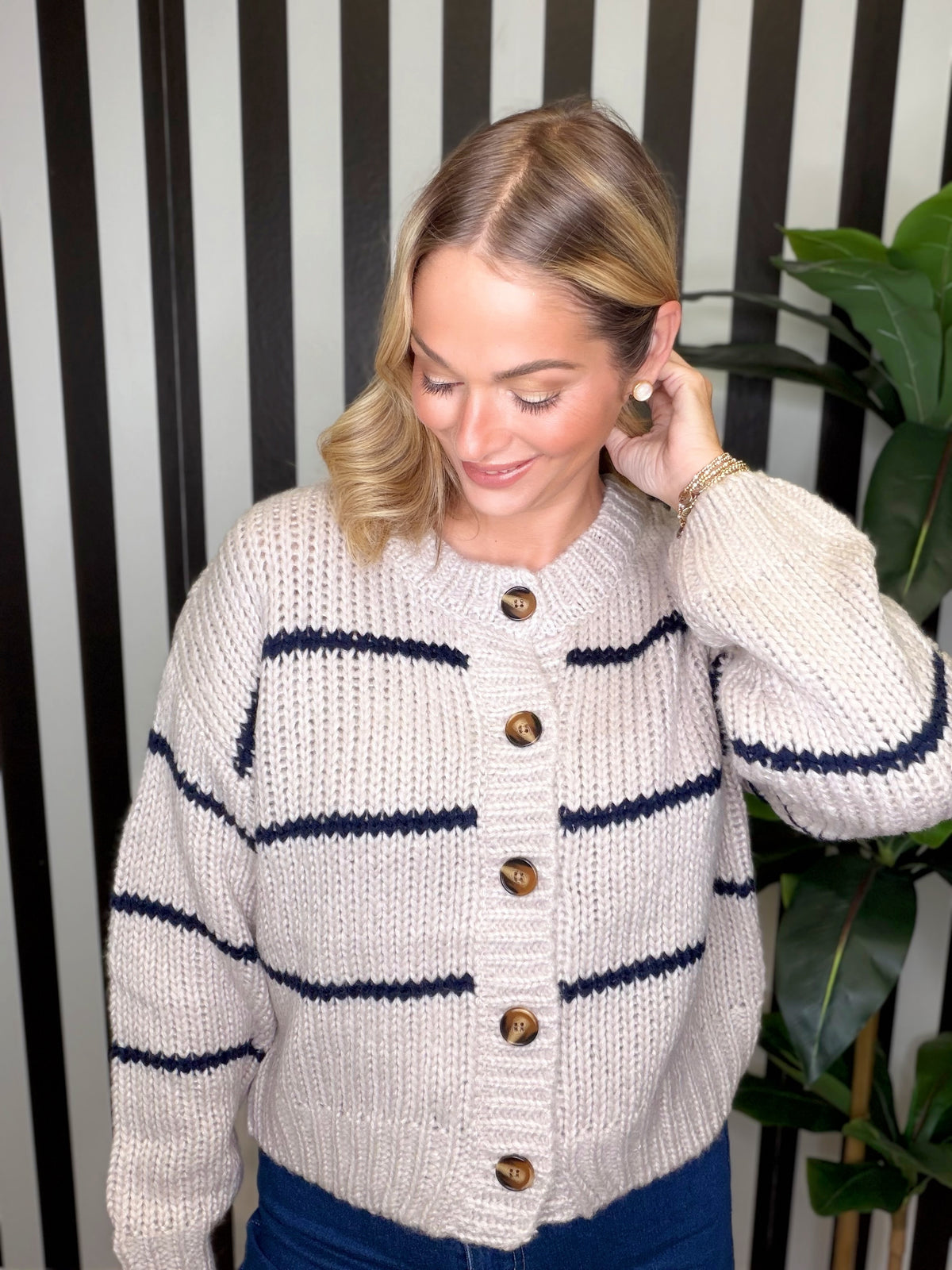 A woman wearing a cream-colored chunky knit cardigan with navy blue horizontal stripes and brown buttons, standing in front of a black and white striped wall with a green plant in the background.