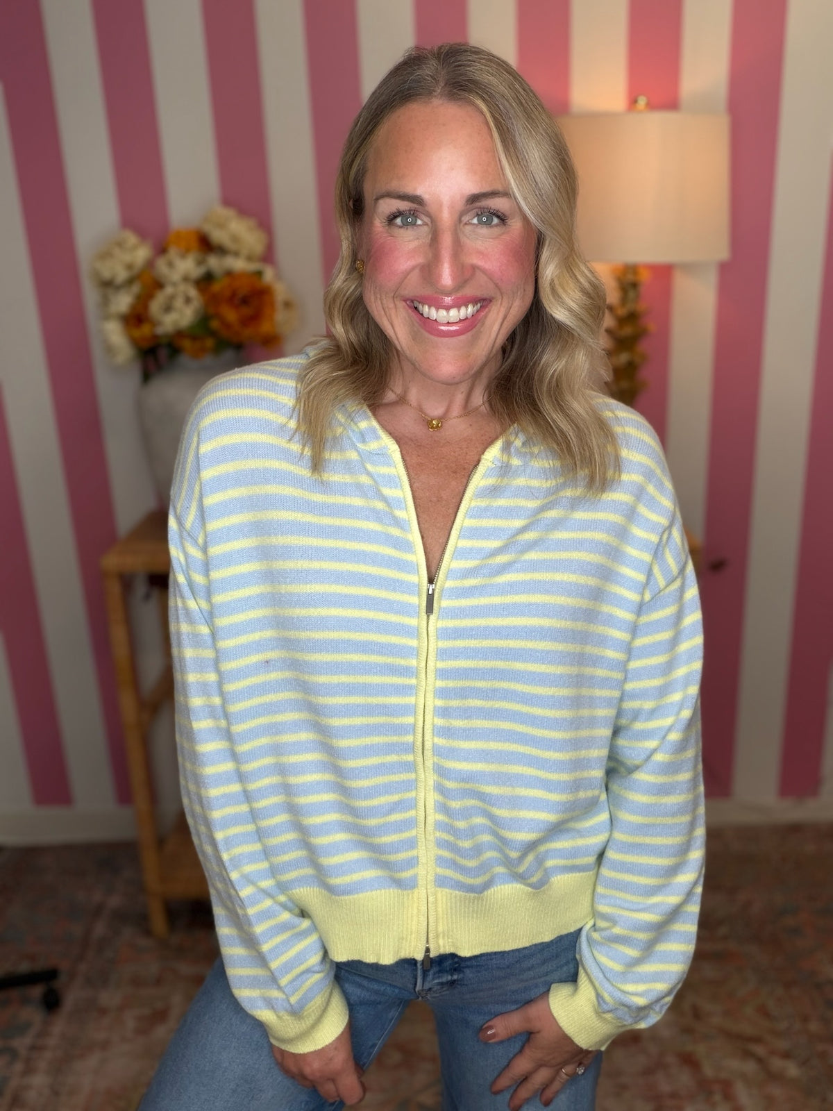 A smiling blonde woman wearing a pastel yellow and light blue striped zip-up cardigan with a yellow zipper, ribbed collar, cuffs, and hem, paired with blue jeans, posed in front of pink and white striped wallpaper.