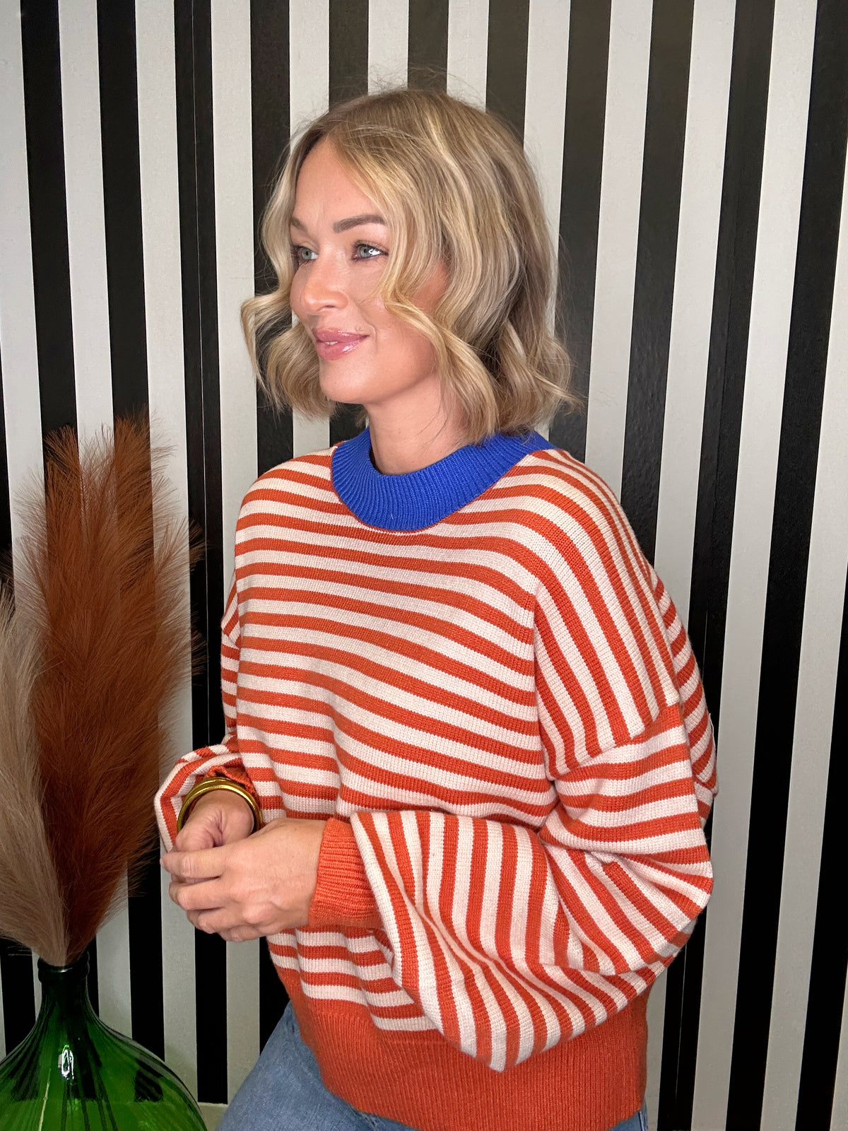 A woman wearing a vibrant orange and white striped knit sweater with a contrasting royal blue crew neck, standing in front of a black and white striped wall.