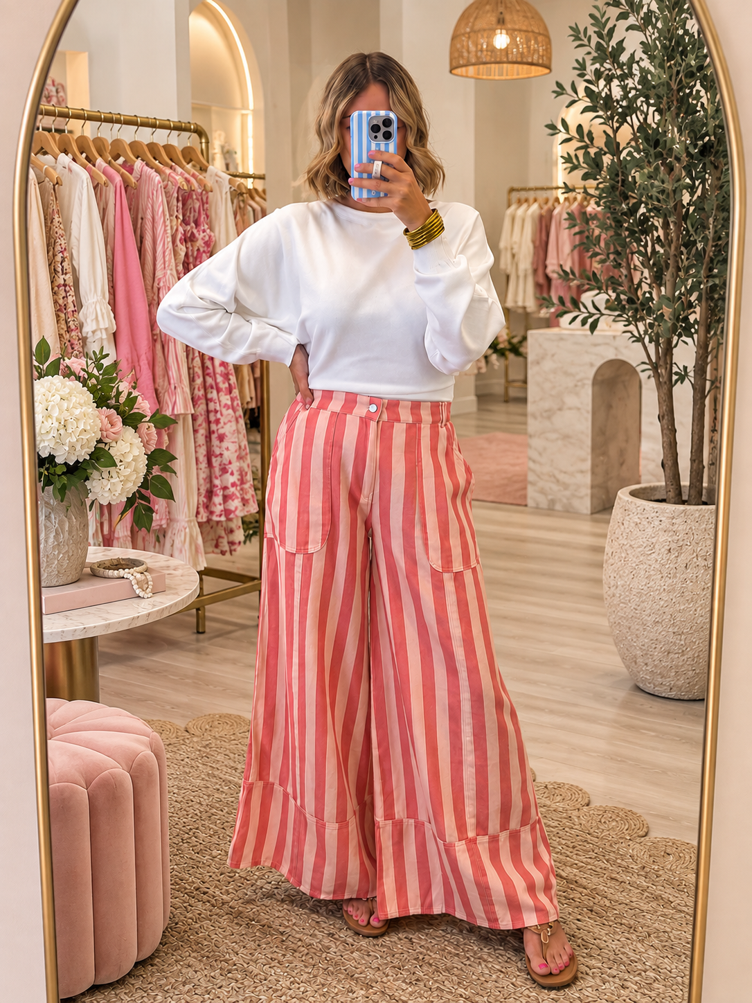 A woman takes a mirror selfie in a boutique, wearing a white long-sleeve top and high-waisted, wide-leg trousers with vertical pink and white stripes. The boutique features a gold-framed mirror, a pink velvet ottoman, and racks of clothing in the background.