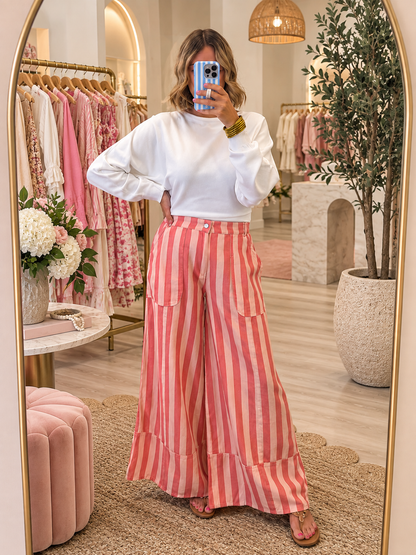 A woman takes a mirror selfie in a boutique, wearing a white long-sleeve top and high-waisted, wide-leg trousers with vertical pink and white stripes. The boutique features a gold-framed mirror, a pink velvet ottoman, and racks of clothing in the background.