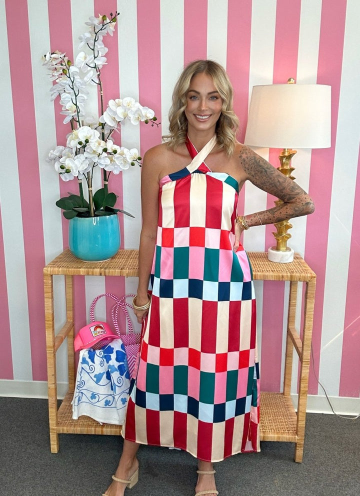 A woman modeling a colorful halter-neck midi dress with a red, pink, blue, and white checkered pattern, standing in front of a pink and white striped wall.