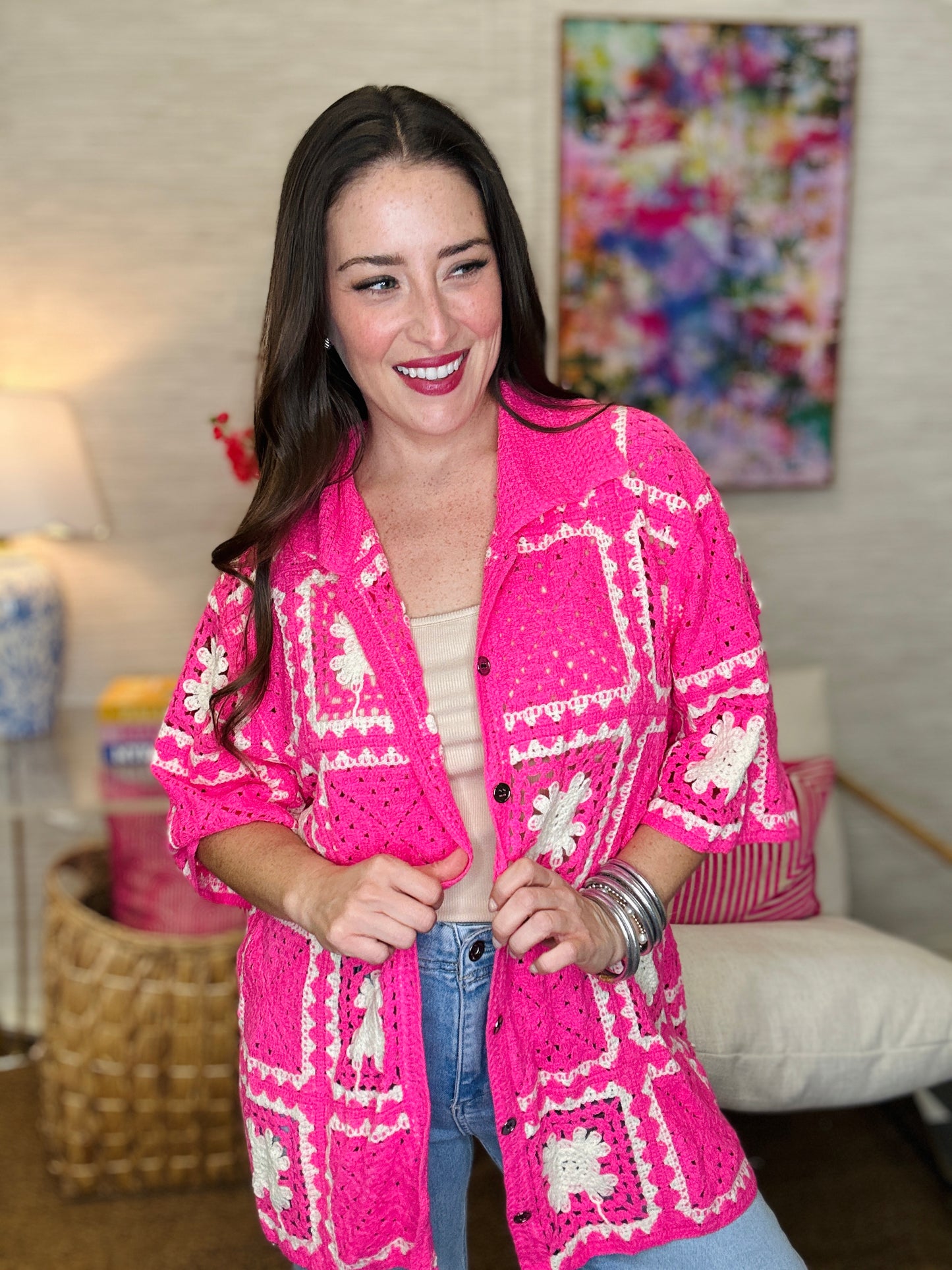 Woman wearing a bright pink crochet cardigan with white patterns in a living room setting.