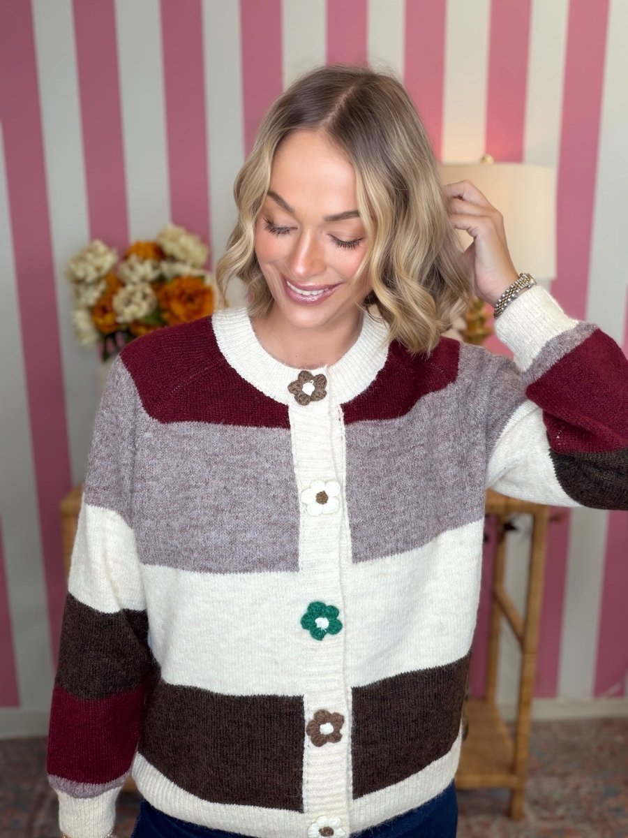 A woman wearing a chunky knit cardigan sweater with horizontal stripes in cream, taupe, burgundy, and dark brown, featuring decorative flower-shaped buttons. She is standing in front of a pink and white striped wall.