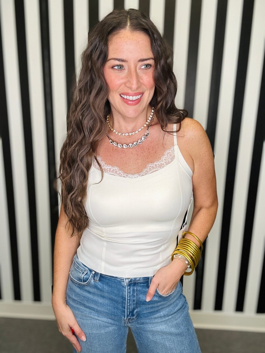A woman with long wavy brown hair smiling, wearing a white lace-trimmed camisole tank top and blue jeans, accessorized with a pearl necklace and gold bracelets, standing in front of a black and white striped wall.