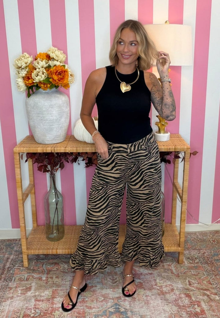 A woman stands in a room with pink and white striped wallpaper, wearing a black sleeveless top and wide-leg zebra-print pants. She is posing in front of a wooden console table decorated with a large white vase of flowers and a lamp.