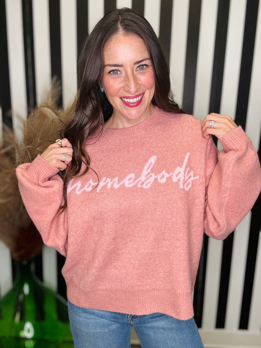 A woman wearing a dusty pink crewneck sweater with the word 'homebody' in white script across the chest, paired with blue jeans, posing in front of a black and white striped background.