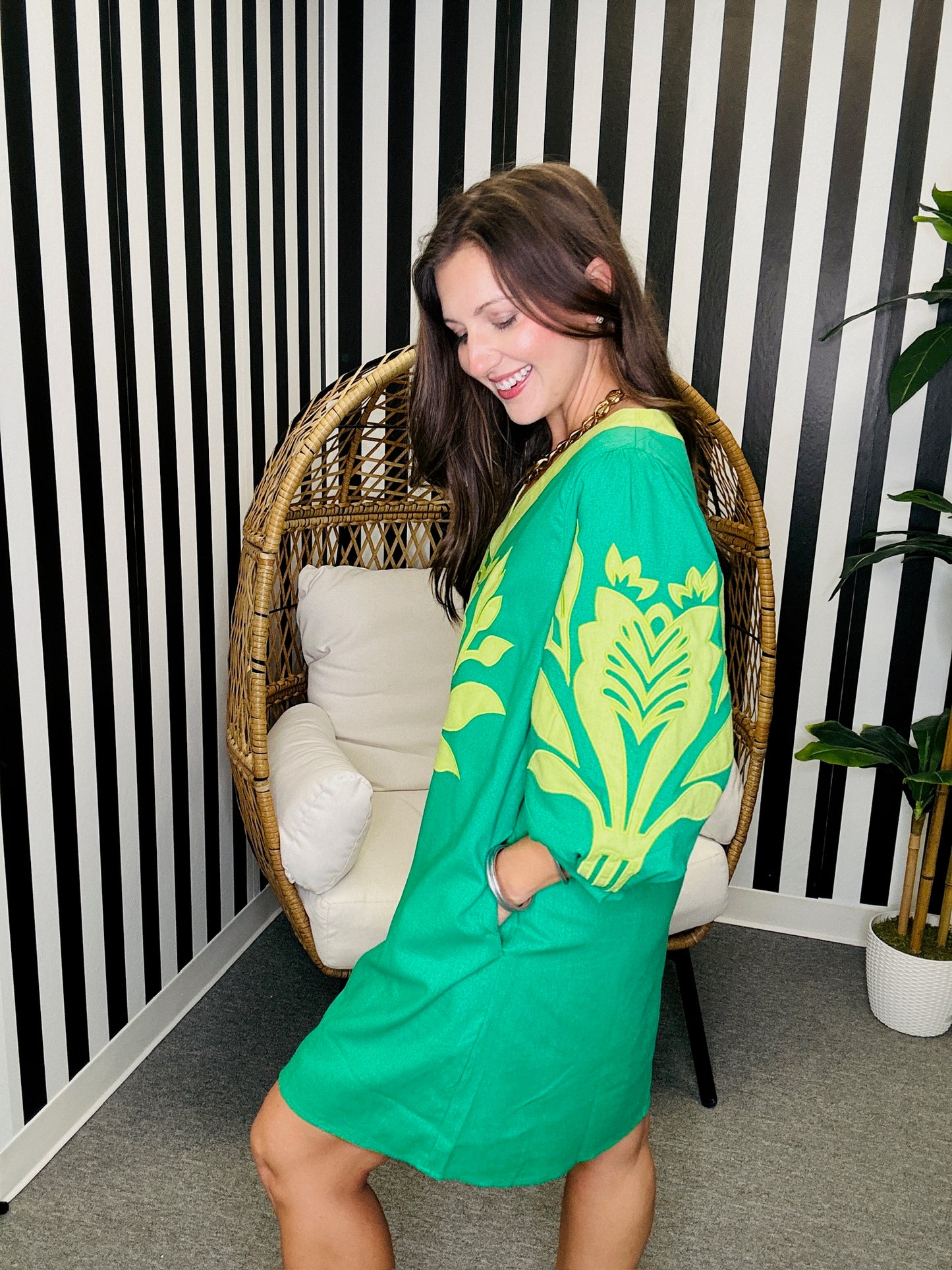 Woman wearing a green dress with yellow patterns sitting on a wicker chair against a black and white striped wall.