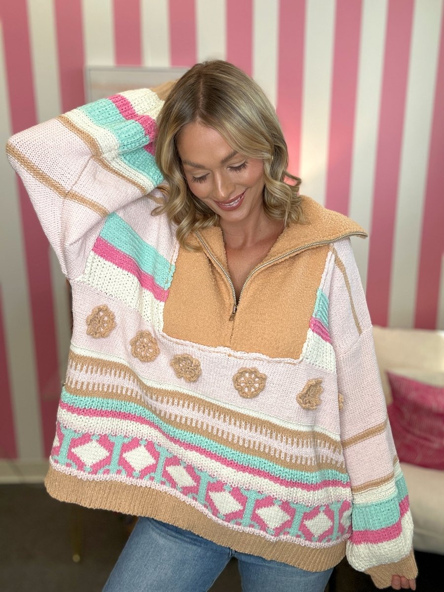A woman wearing a chunky knit half-zip sweater with a tan fleece collar, featuring horizontal stripes in pink, mint green, white, and beige, posed against a pink and white striped wall.