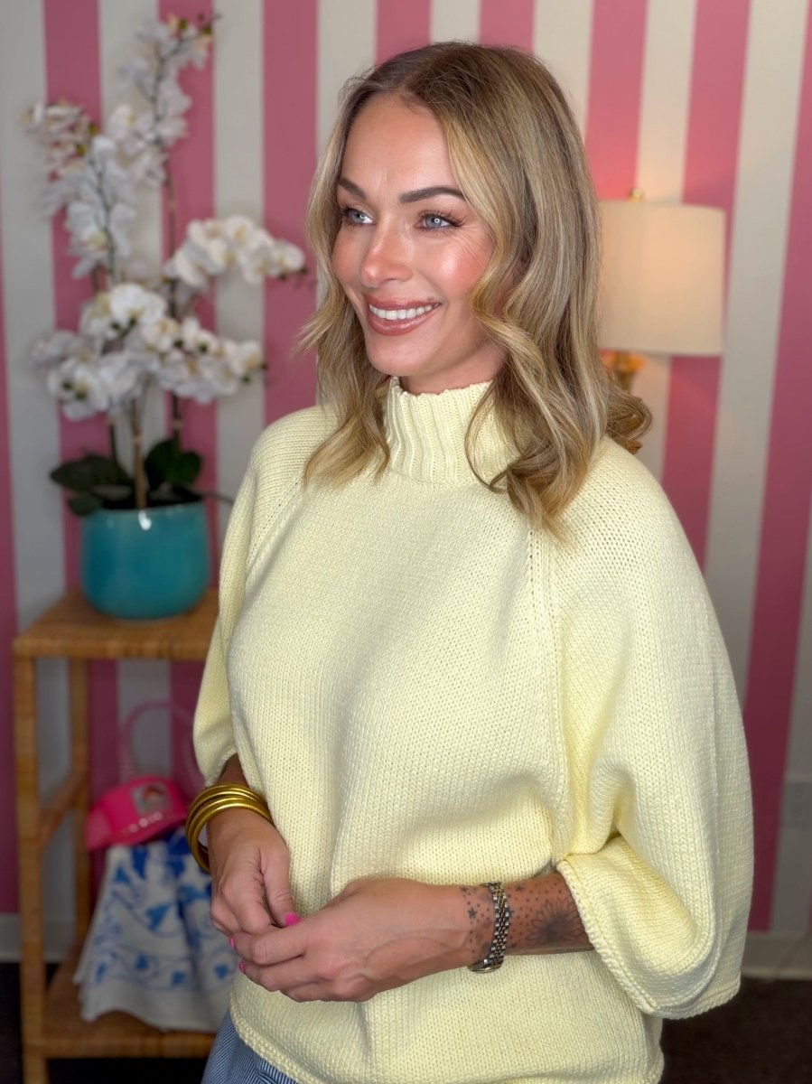 A smiling blonde woman wearing a pale yellow knit mock-neck sweater, standing in front of a pink and white striped wall with a side table and flowers in the background.