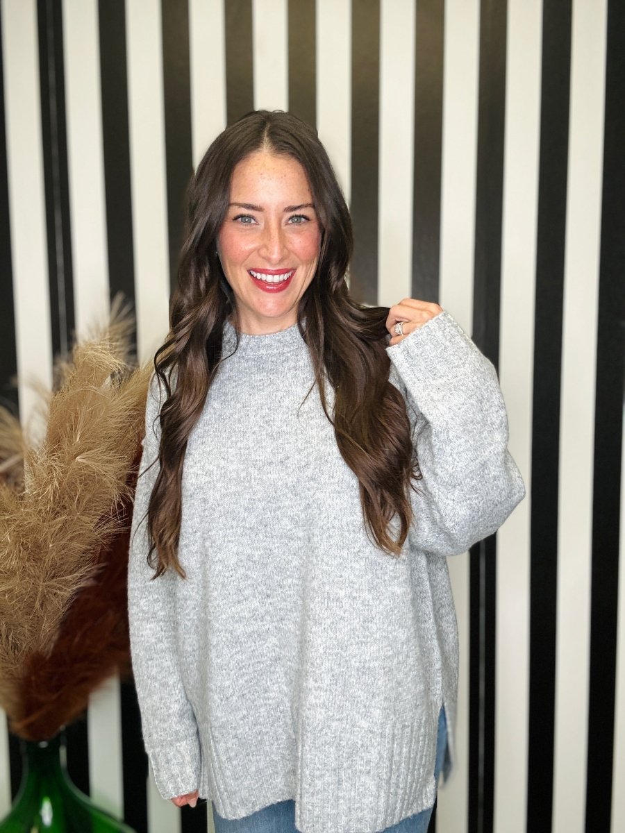 A smiling woman with long brown hair wearing a light gray heathered knit sweater, standing in front of a black and white striped wall with dried grass decor on the left.