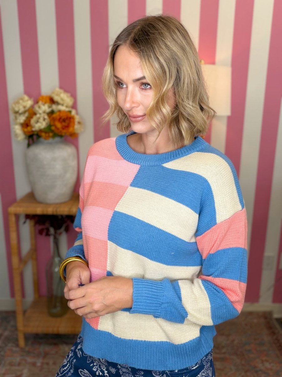 A woman wearing a colorful striped knit sweater with blocks of blue, pink, and cream, standing in front of a pink and white striped wall.