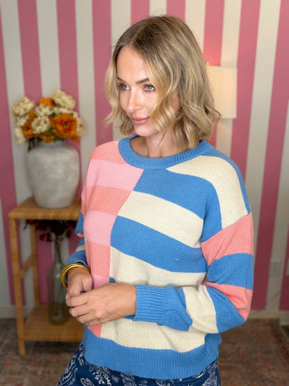 A woman wearing a colorful striped knit sweater with blocks of blue, pink, and cream, standing in front of a pink and white striped wall.