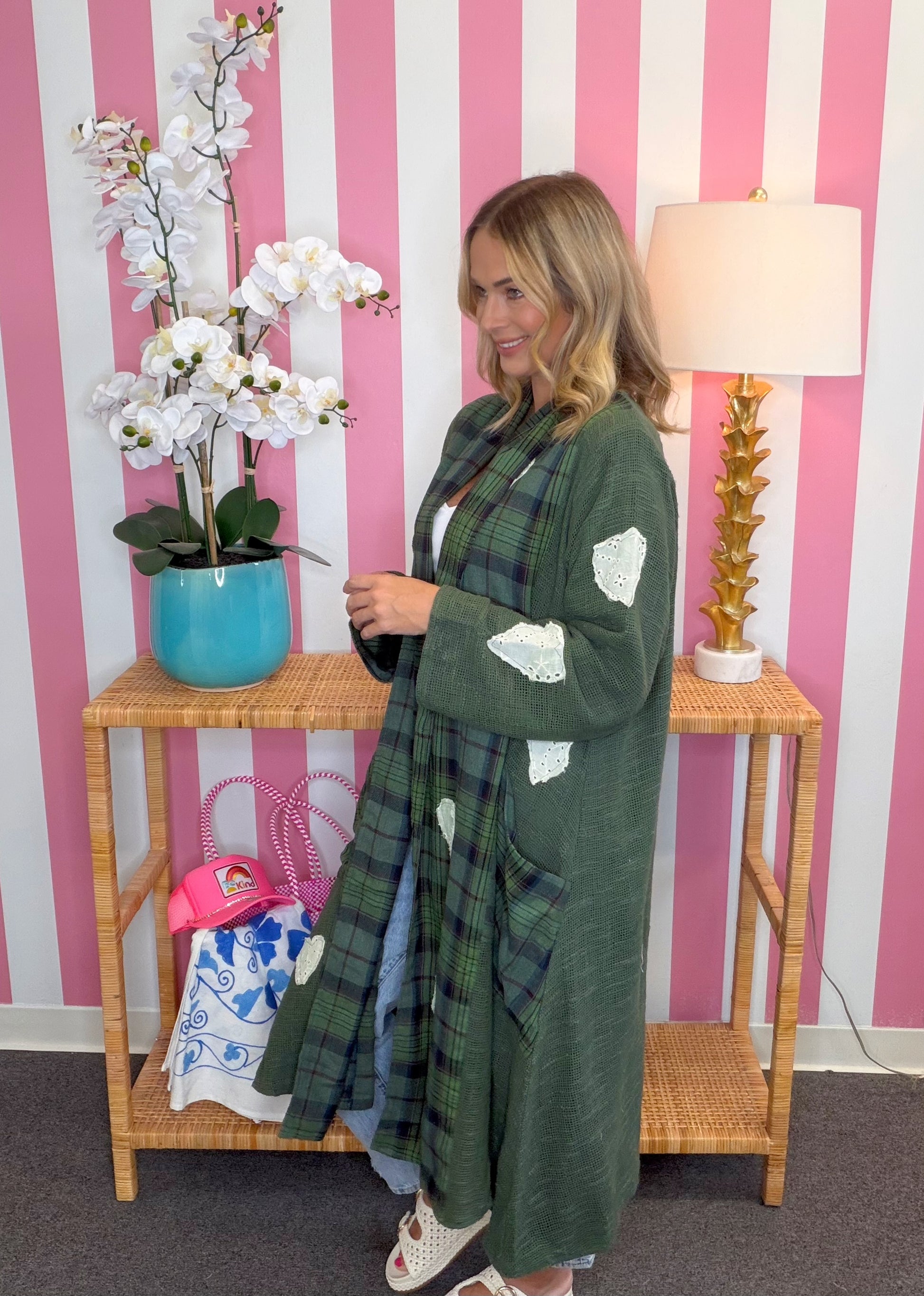 A woman wearing a long green plaid cardigan with white patchwork details stands in front of a pink and white striped wall, next to a wooden console table with a blue orchid pot and a lamp.