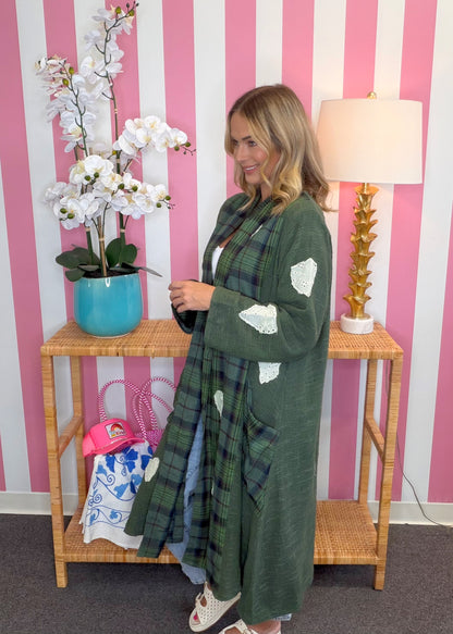 A woman wearing a long green plaid cardigan with white patchwork details stands in front of a pink and white striped wall, next to a wooden console table with a blue orchid pot and a lamp.