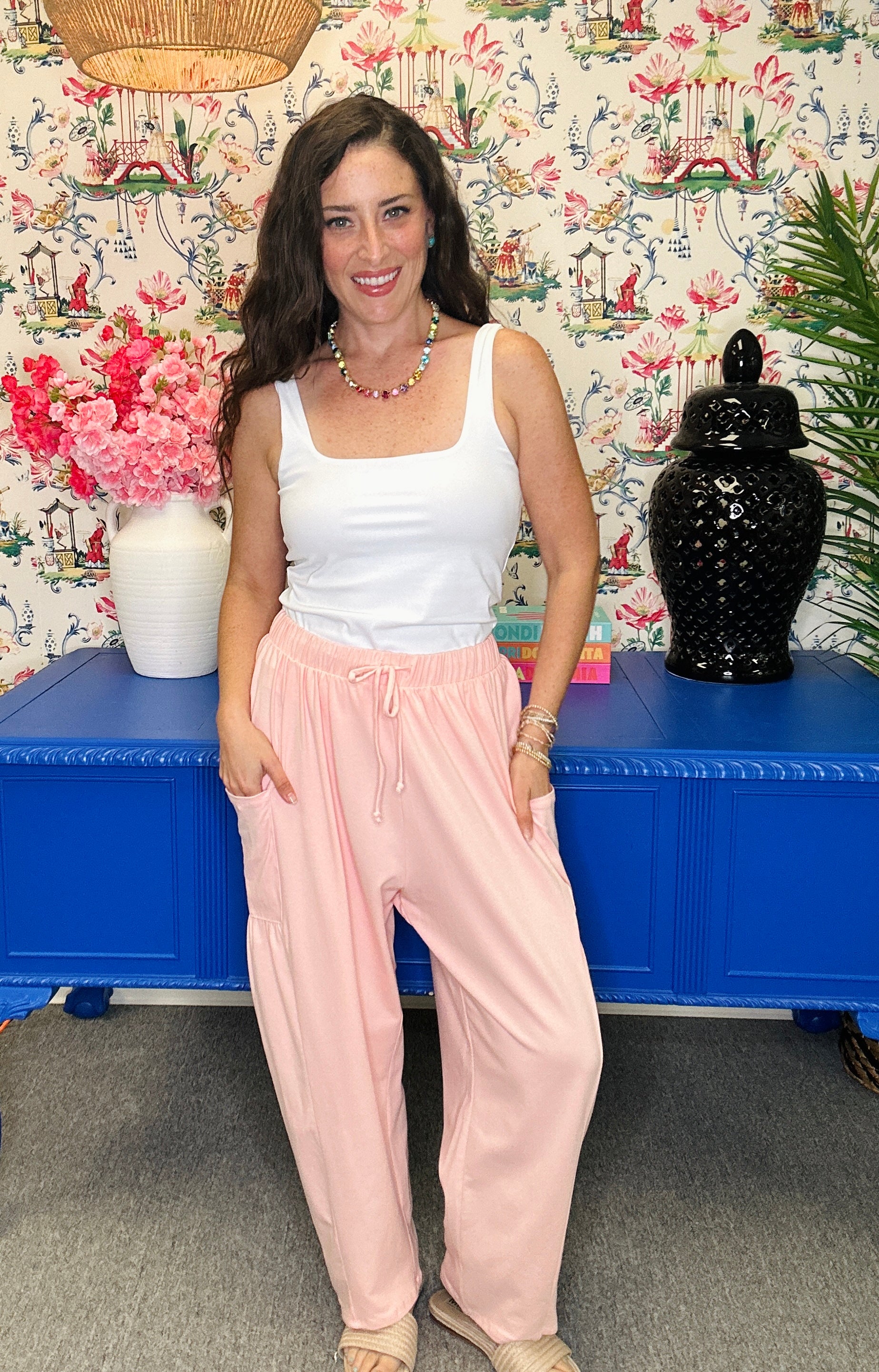 A woman posing in a white tank top and light pink drawstring sweatpants, standing in front of a blue cabinet and a floral wallpaper background.