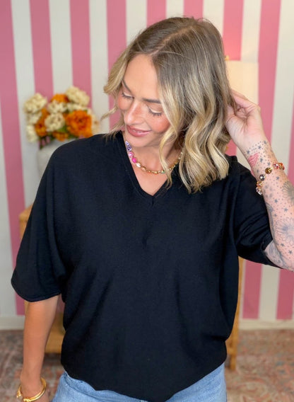 A woman wearing a black short-sleeve V-neck top and light blue jeans, standing in front of a pink and white striped wall with flowers in the background.