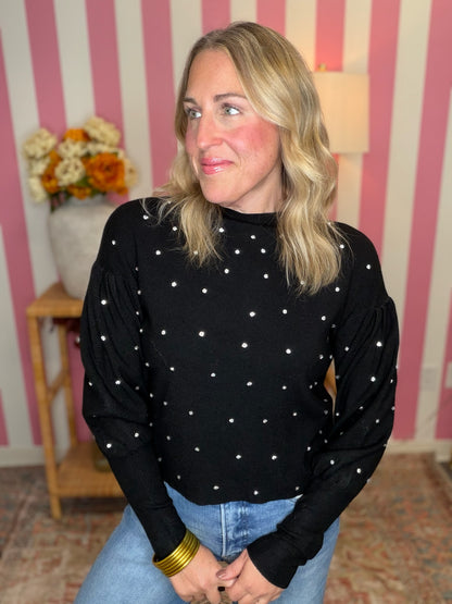 A woman with blonde hair wearing a black long-sleeve mock neck top with white polka dots, paired with blue jeans and a gold bracelet, posing in front of a pink and white striped wall.