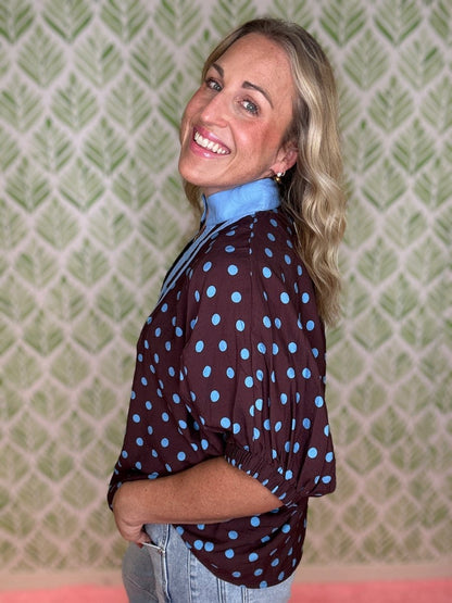 A smiling blonde woman wearing a dark brown long-sleeve blouse with light blue polka dots and a light blue collar, paired with light-wash jeans, standing in front of a green and white patterned wallpaper background.