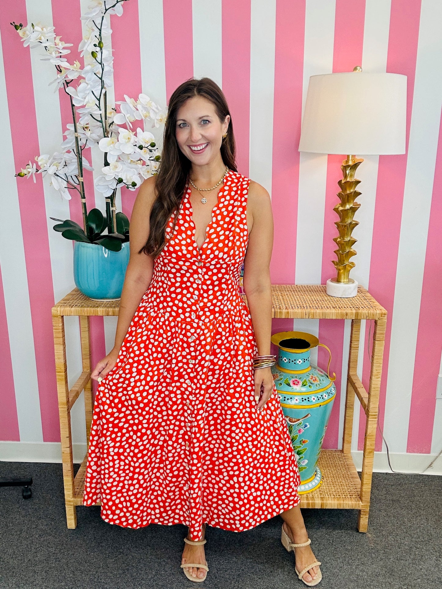 Woman in a red floral dress standing in front of a pink and white striped wall with decorative vases and a lamp.
