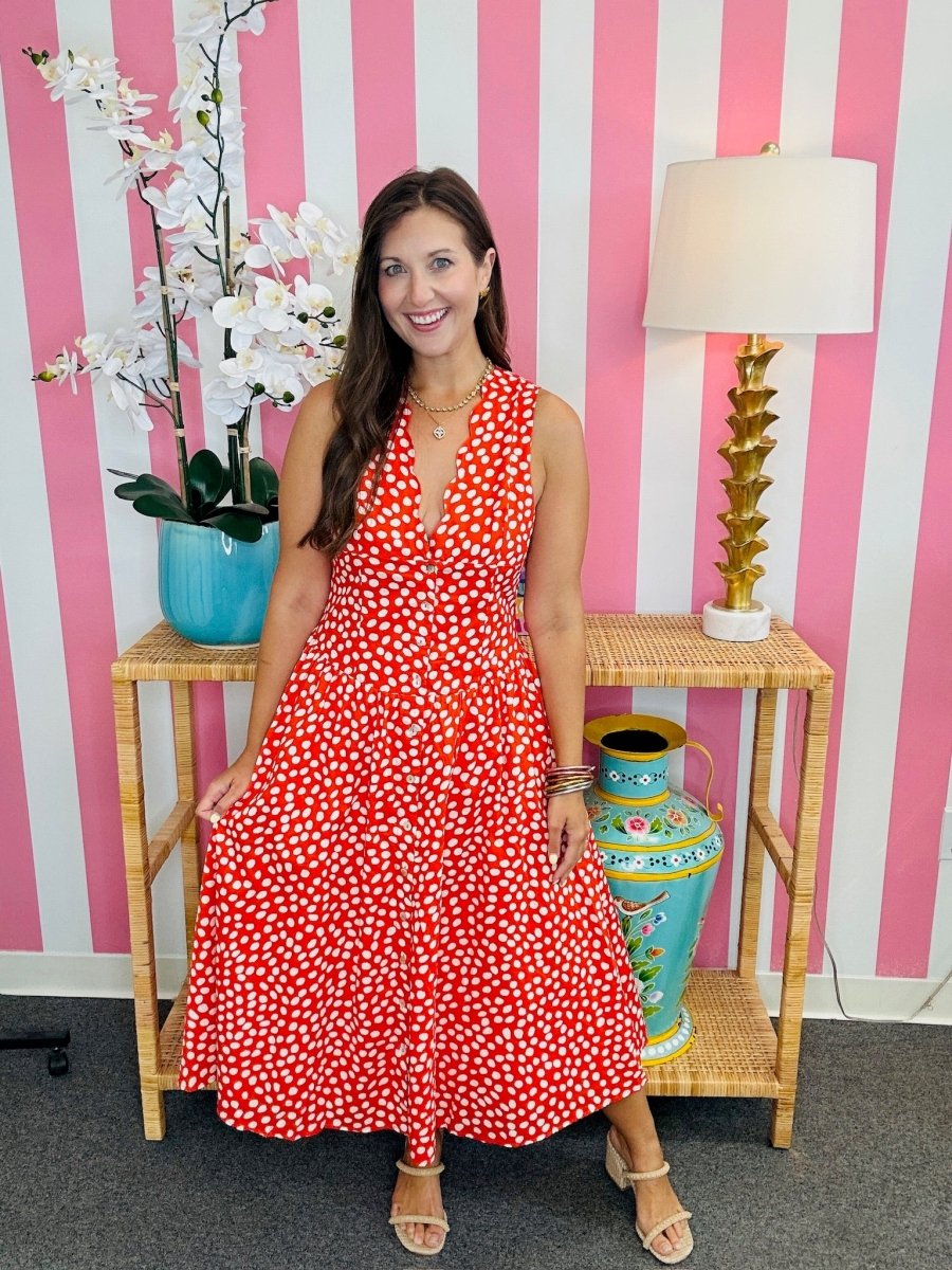 Woman in a red floral dress standing in front of a pink and white striped wall with decorative vases and a lamp.