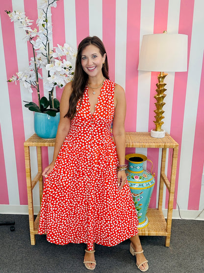 Woman in a red floral dress standing in front of a pink and white striped wall with decorative vases and a lamp.