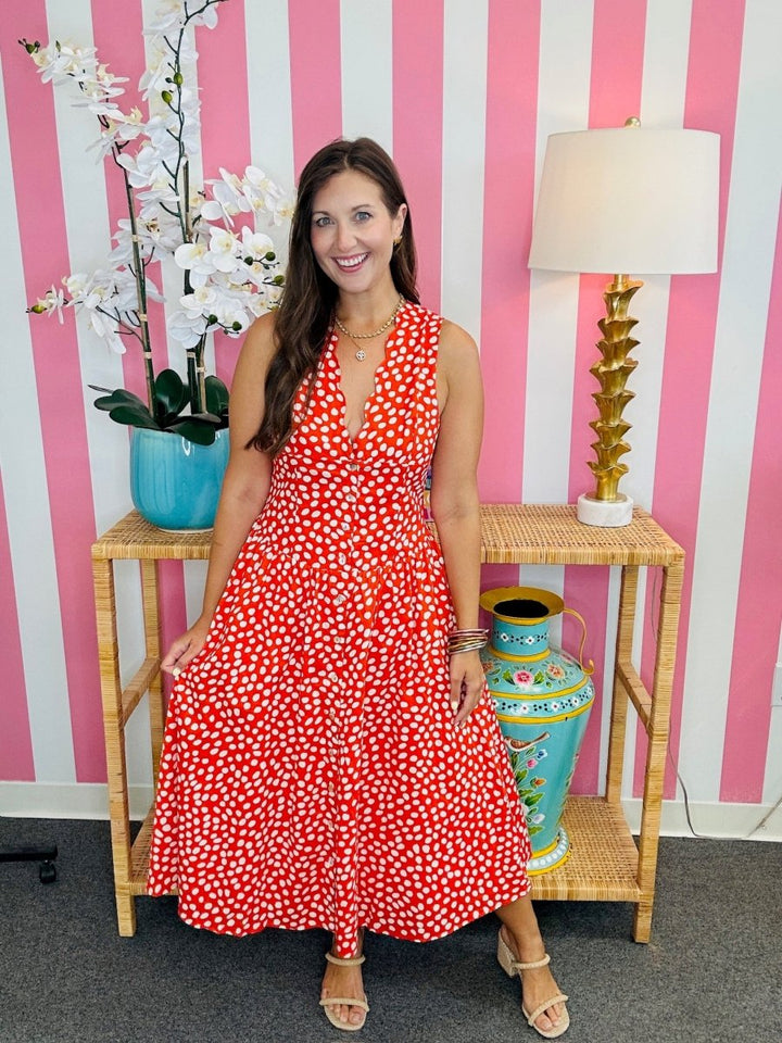 Woman in a red floral dress standing in front of a pink and white striped wall with decorative vases and a lamp.