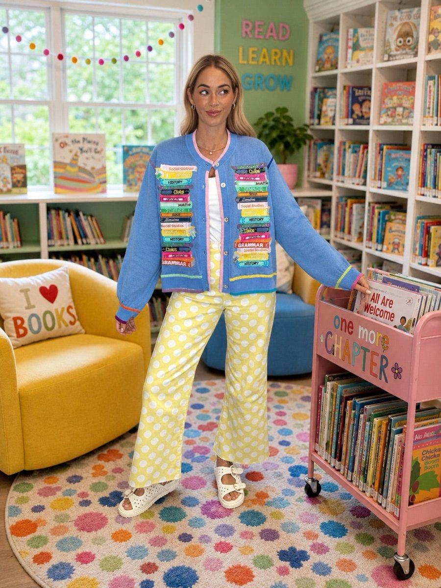 A woman stands in a colorful children's bookroom wearing a light blue cardigan with book-cover panels, yellow-and-white polka dot pants, and white slide sandals. She is posed beside a pink book cart and bookshelves.