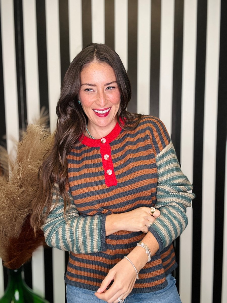 A woman smiling and wearing a striped knit henley sweater with a red collar and buttons, layered over a green and beige striped long-sleeve shirt, set against a black and white striped background.