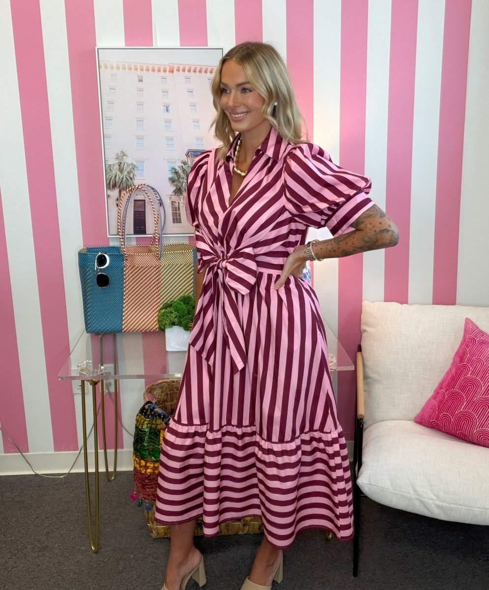 A woman models a pink and white vertically striped midi dress with puff sleeves and a tie waist, standing in front of a pink and white striped wall.