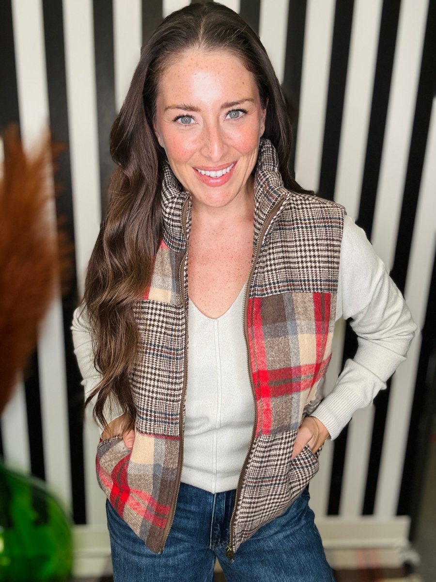 A woman wearing a sleeveless zip-front vest with a plaid pattern in brown, beige, and red, layered over a white long-sleeve top and blue jeans, standing against a black and white striped background.