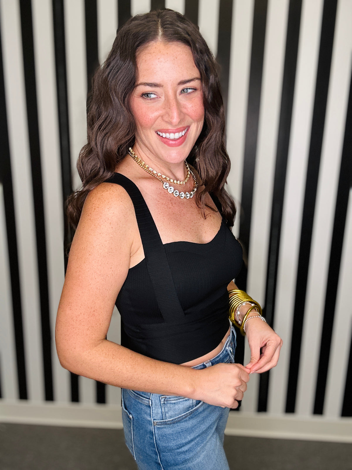 A smiling woman with long wavy brown hair wears a black sleeveless crop top and blue jeans, accessorized with layered gold necklaces and bracelets, standing in front of a black and white vertically striped wall.