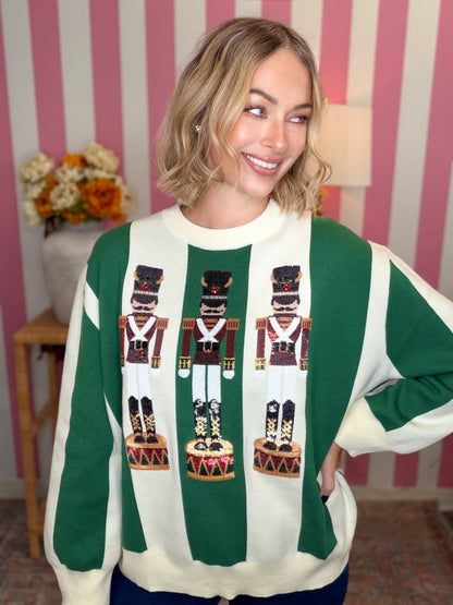 A woman wearing a green and cream striped crewneck sweater with three nutcracker figures on the front, posed in front of a pink and white striped wall.