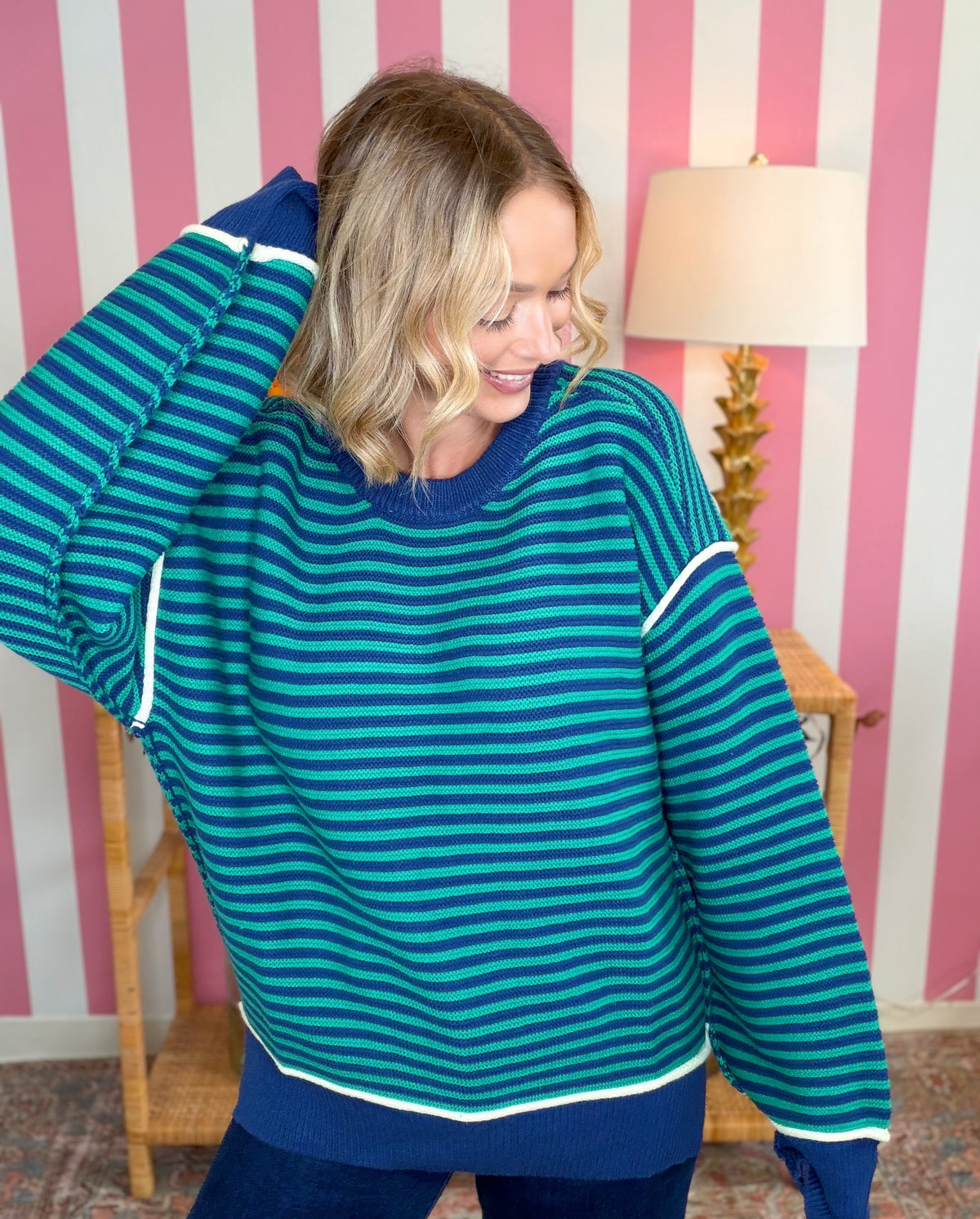 A woman wearing a teal and navy striped sweater with white trim, posing in front of a pink and white striped wall.