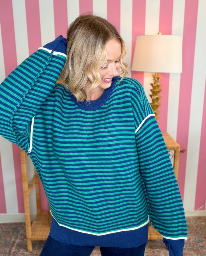 A woman wearing a teal and navy striped sweater with white trim, posing in front of a pink and white striped wall.