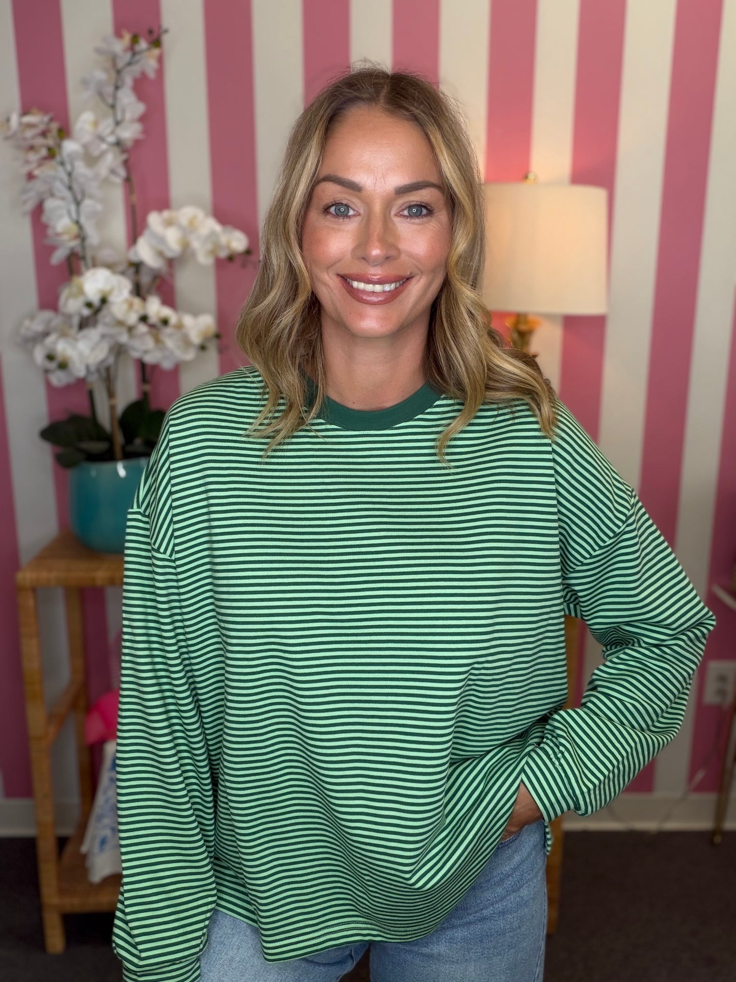 A woman wearing a green and white horizontally striped long-sleeve top, standing in front of a pink and white striped wall with a lamp and flowers in the background.