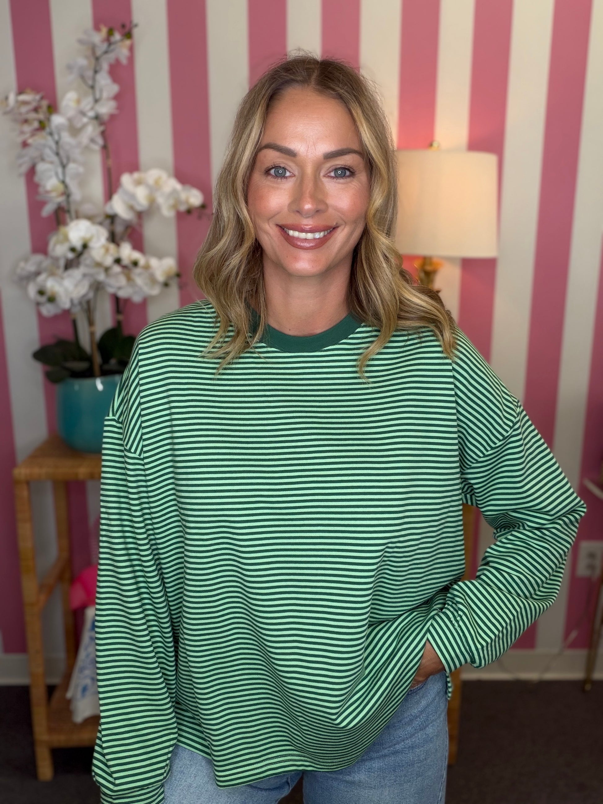 A woman wearing a green and white horizontally striped long-sleeve top, standing in front of a pink and white striped wall with a lamp and flowers in the background.