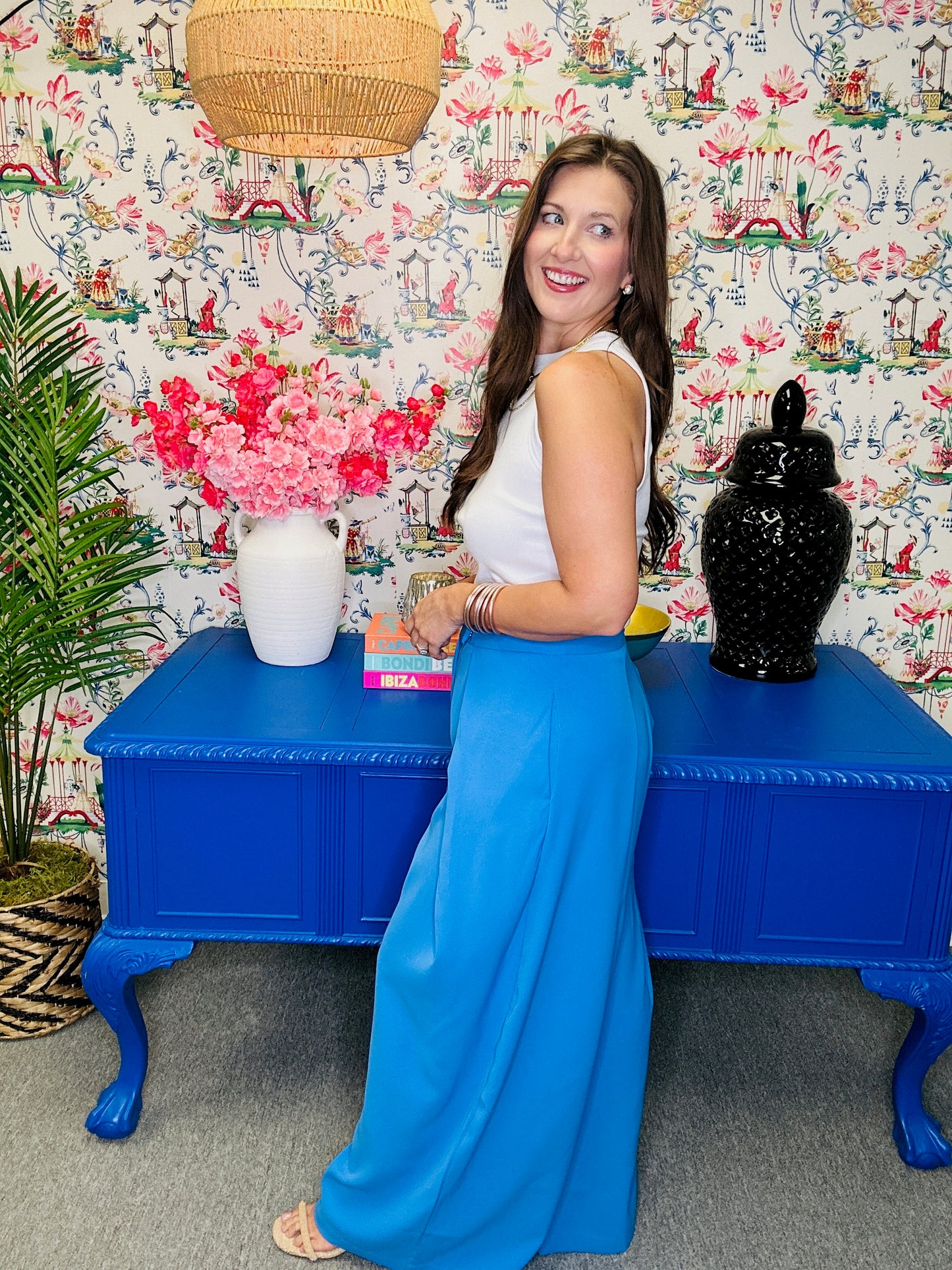 Woman in a blue dress standing next to a blue table with decorative items against a patterned wall.