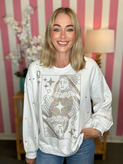 A smiling blonde woman wearing a white crewneck sweatshirt with a large gold glittery playing card graphic on the front, set against a pink and white striped wall background.