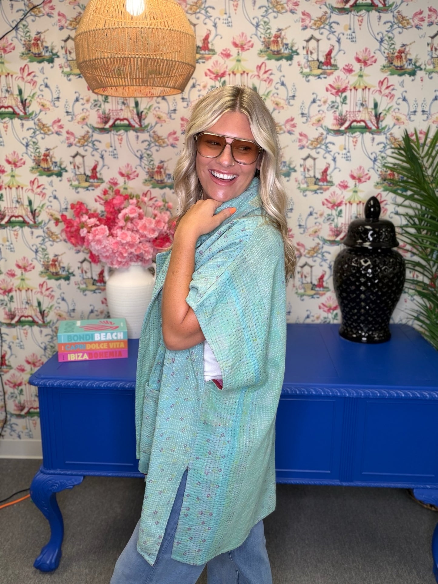 A woman wearing a light mint green patterned shawl over a white top and blue jeans, standing in front of a blue console table and floral wallpaper.