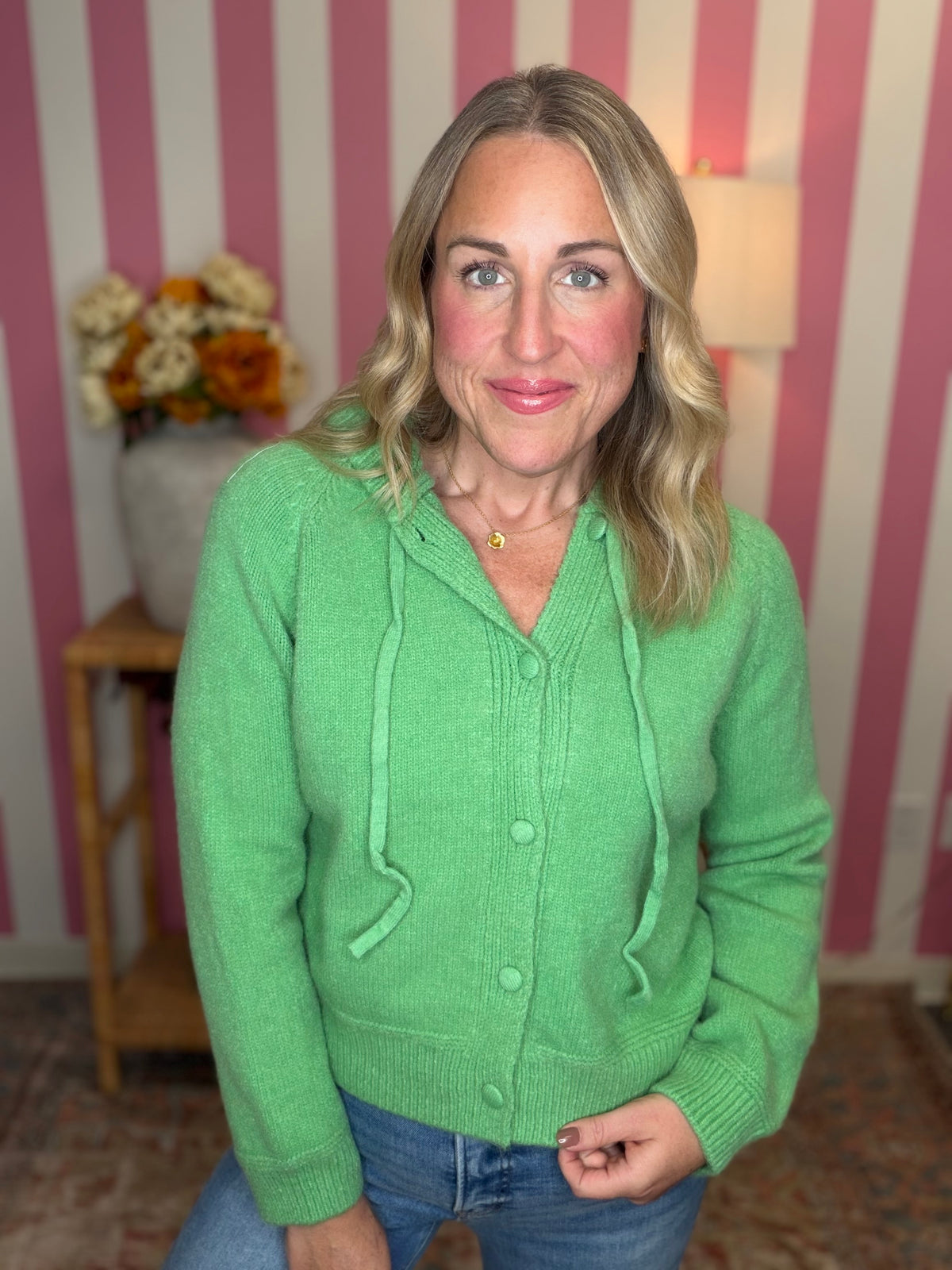 A woman wearing a bright green knit cardigan with a drawstring hood and front buttons, paired with blue jeans, standing in front of a pink and white striped wall.